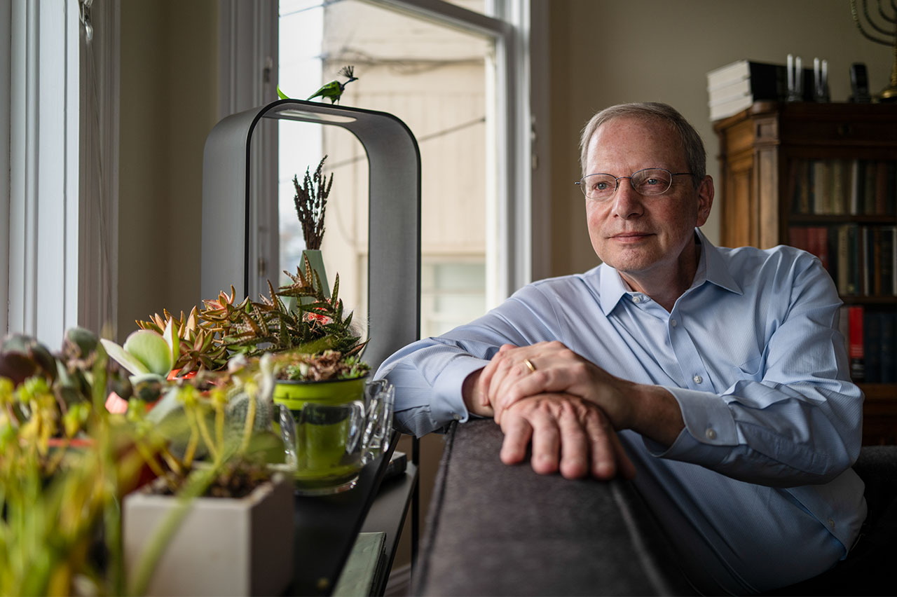 Robert Wachter sits on his couch in his home, resting is arm on the back with succulent pants in the foreground by the window.