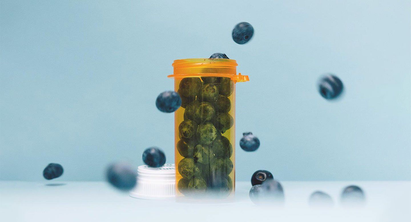 Photo of blueberries falling into a prescription pill bottle in front of a blank blue background.