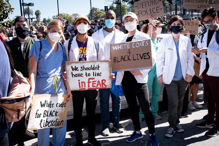 UCSF students at a protest for the killing of George Floyd. They wear scrubs, white coats, and face masks and hold signs that read “White coats for Black liberation,” “We shouldn't have to live in fear,” “White Coast 4 Black Lives,”  and “Defund Police.”