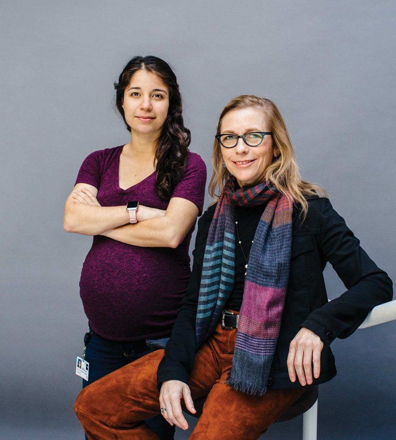 Portrait of Judith Hellman (sitting) and Samira Khakpour Lawton, standing in front of an all grey background.