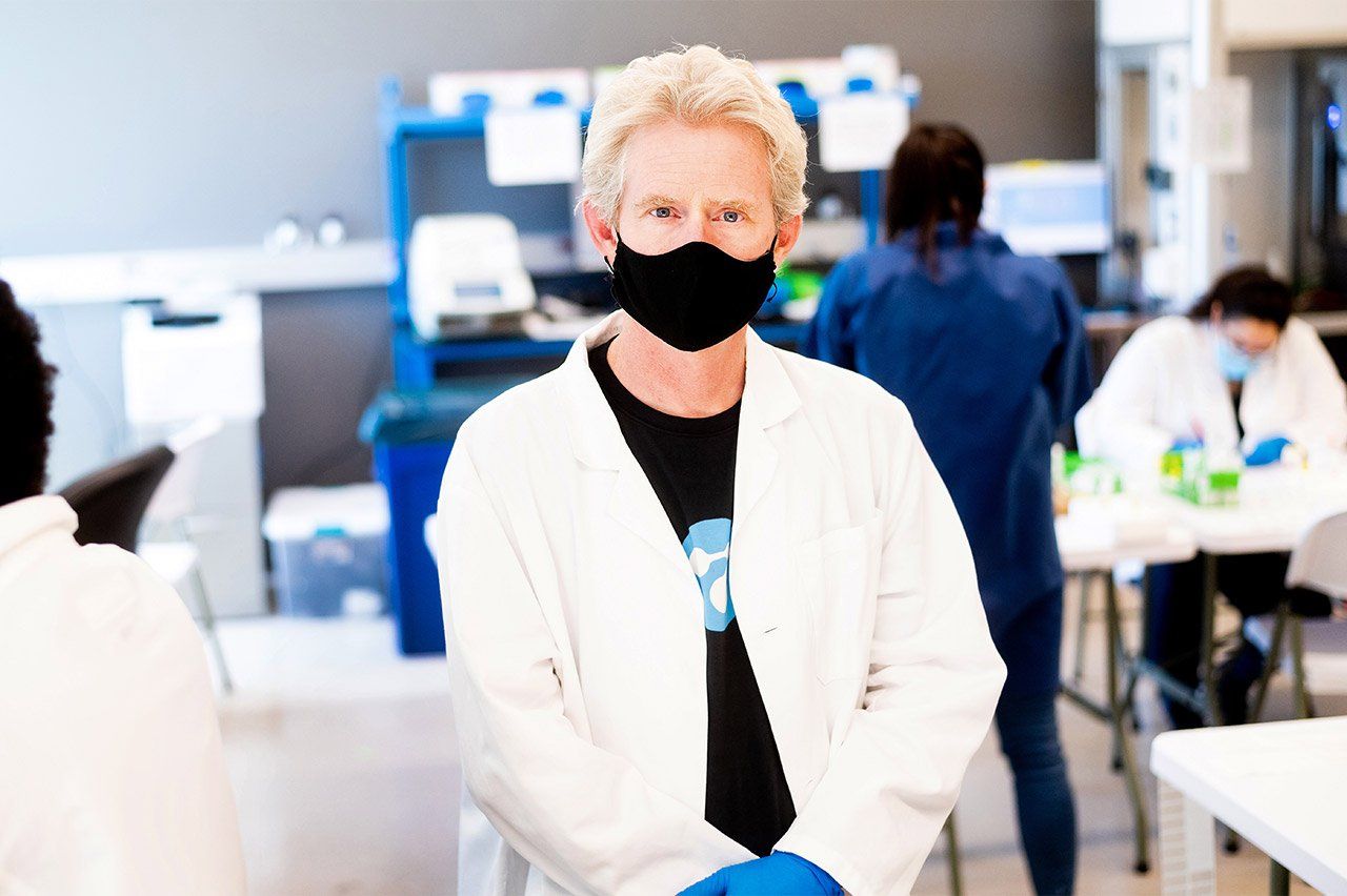 Joe Derisi wearing a black face mask, white lab coat, and blue gloves in the lab; lab team members work behind him.