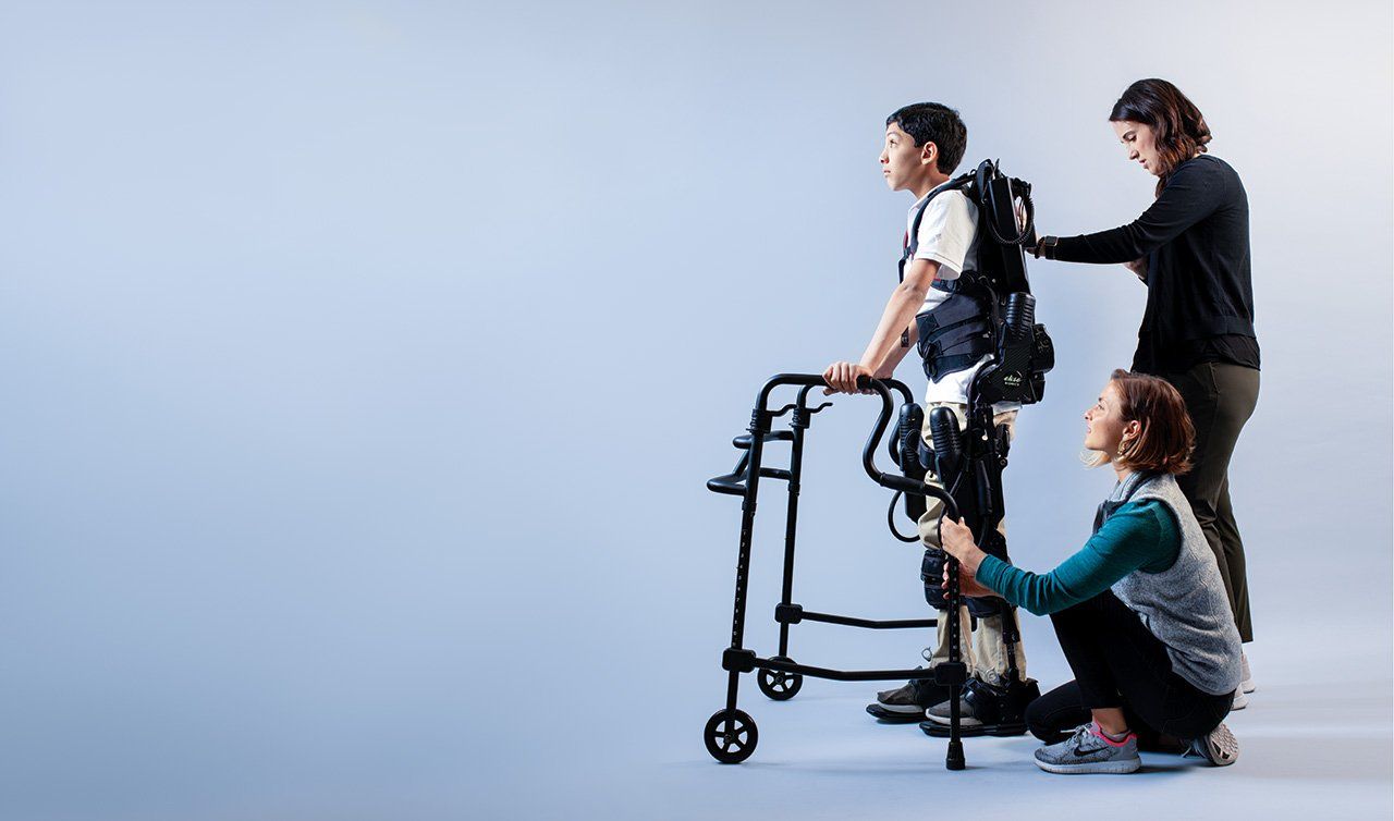 Photo of physical therapist Carolyn Celio, DPT (standing), and rehabilitation aide Cassandra Conlin (kneeling) help Dilan pilot a robotic exoskeleton in front a blank grey background.