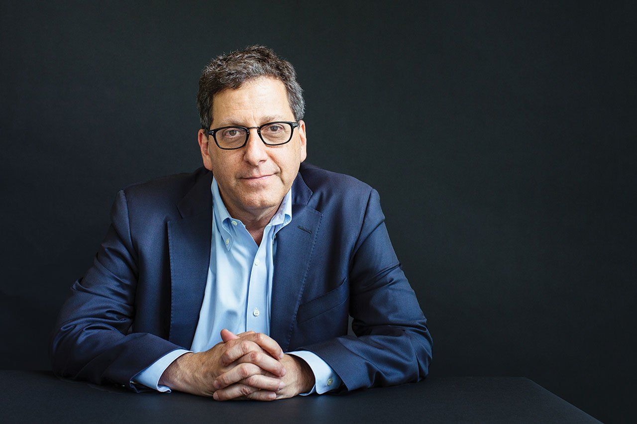 Portrait of Matthew State, MD, PhD, sitting at a black table with his hands folded, resting on the table, in front of a blank black background.