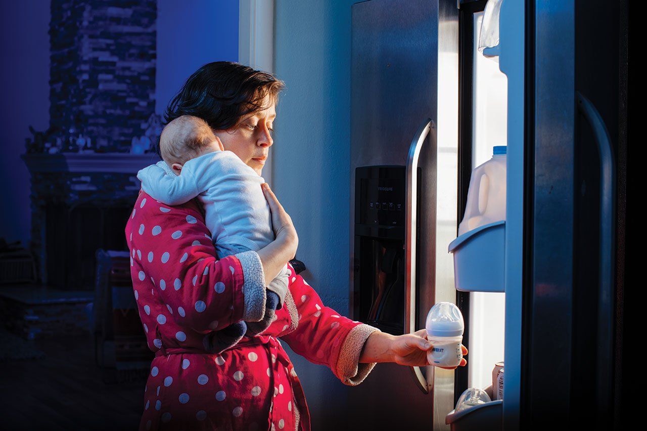 Photo of a tired mother with a newborn baby, reaching for a bottle of milk from the refrigerator at night.