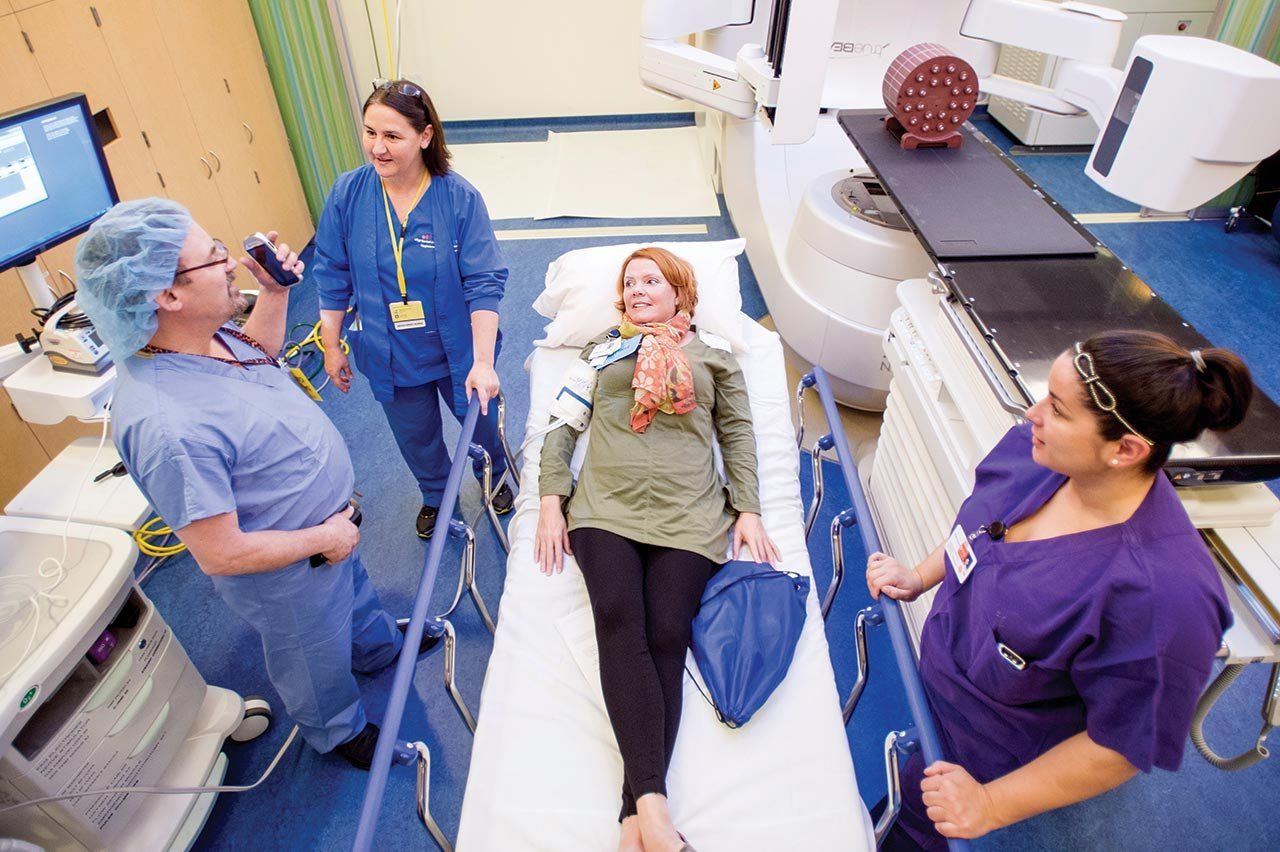 A woman smiles and lays on a hospital gurney; a doctor and nurses stand around her having a discussion. 