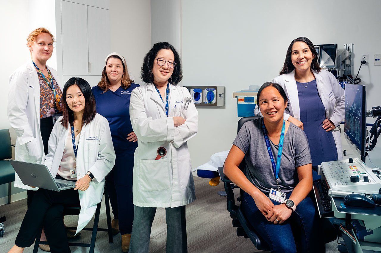 Photo of six women in a medical clinic room. Two women on the left wear white doctor's coats and one holds a computer. Another woman on the left is in blue nurse's scrubs. A woman in the middle wears a white doctor's coat and stands with her arms crossed. On the right side one woman sits at a chair in front of clinical computer device, with images of brain scans on the screen. Another woman in a white doctor's coat stands behind her with her arm resting on the chair.