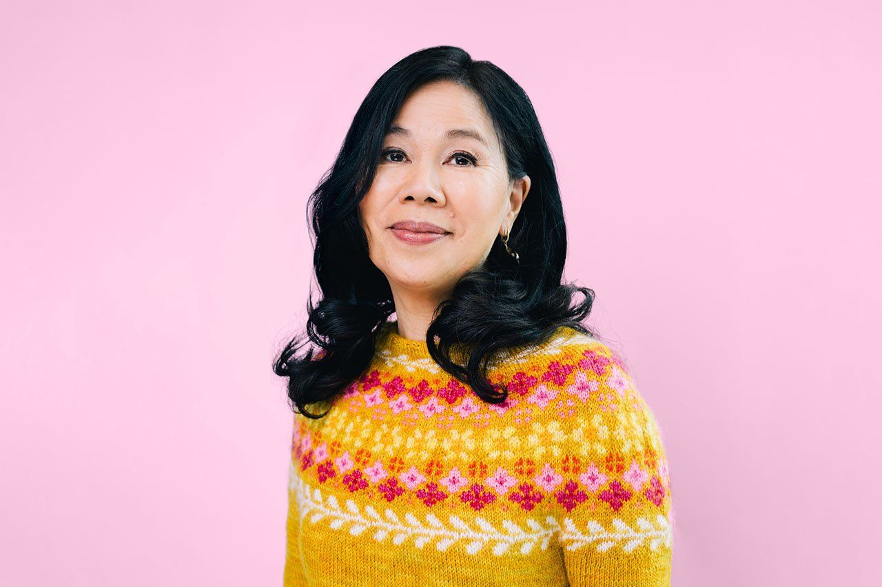 Joanne Chun stands in front of a pink background in a colorful, pink and yellow, hand-knitted, patterned sweater.
