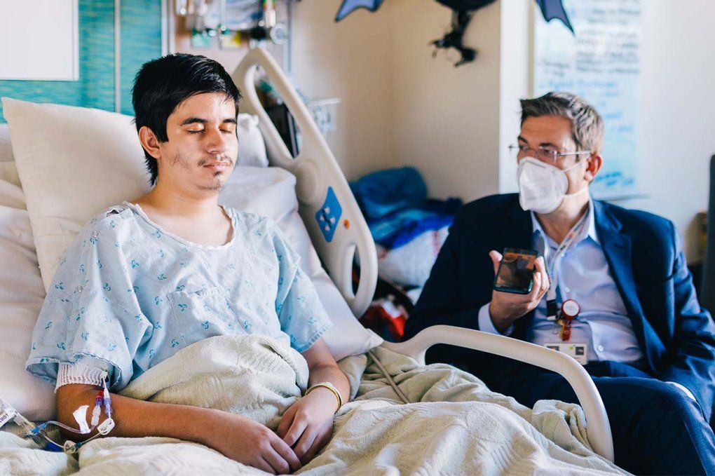 A teenage boy sits in a hospital bed with his eyes closed; Dr. Friedrichsdorf sits next to him.
