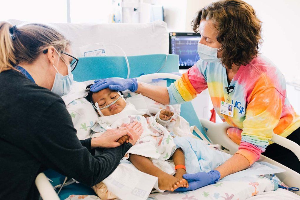 A young child grimaces in a hospital bed as a UCSF message therapist and the boys mother gently touch his feet and belly.