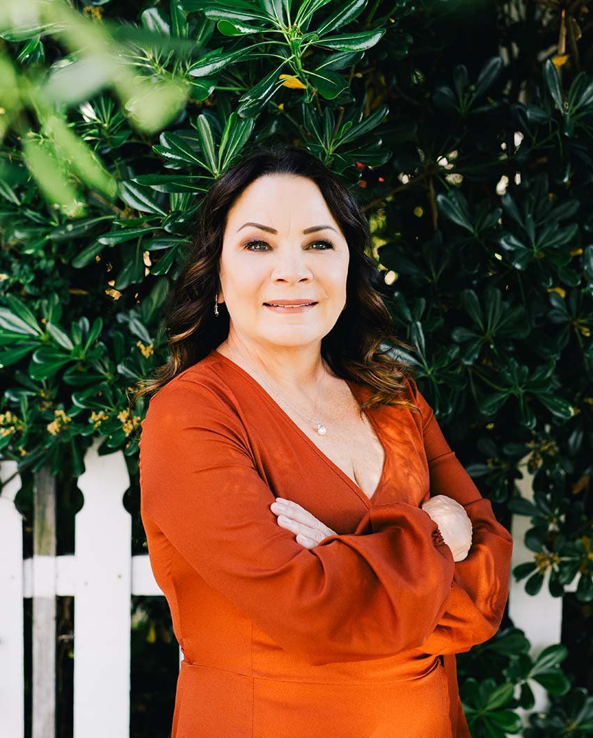 Leeann Bongiorno stands with her arms crossed in front of a green tree and white picket fence.