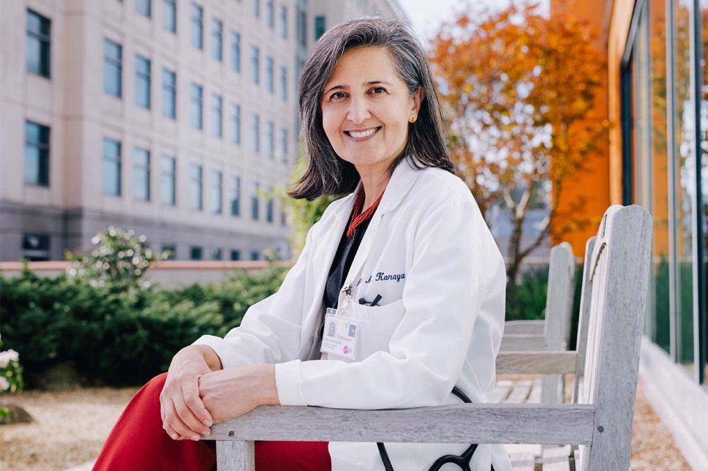 Alka Kanaya wears a white doctor’s coat and sits on a bench in a rooftop garden, with greenery and trees with orange leaves in the background.