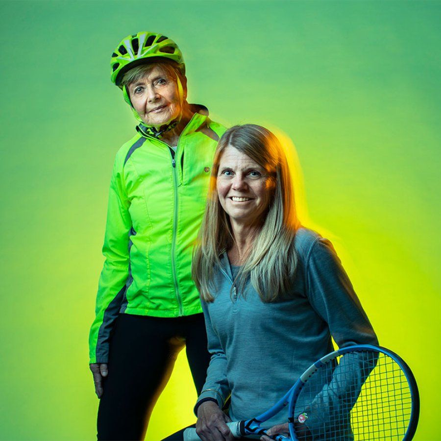 Janice Morgan stands in her cycling gear next to her daughter-in-law, Wendy Morgan, who holds a tennis racket.