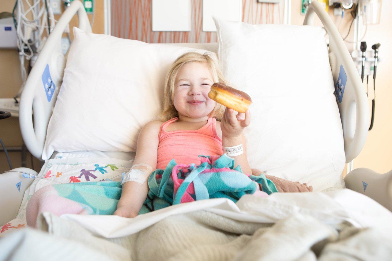 Riley sits in her hospital bed with a donut in her hand and a big smile on her face.