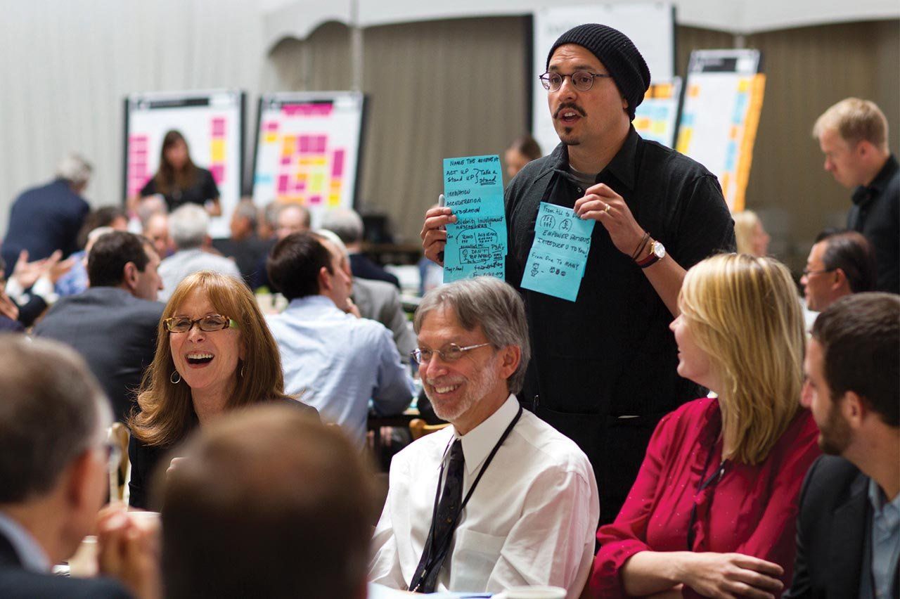 A man holds up some sticky notes and speaks to a table of people at the OME conference.