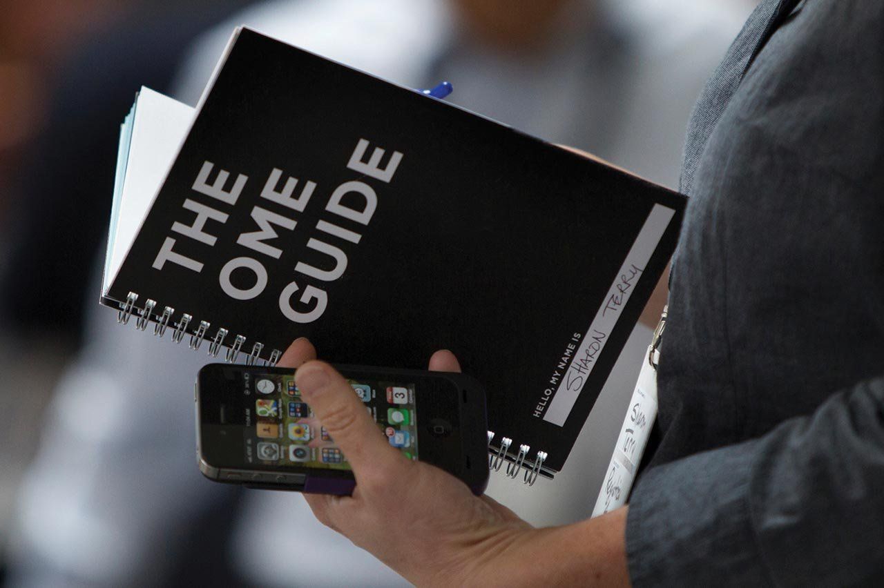 A person holds an iPhone and a notebook that reads "The OME Guide"