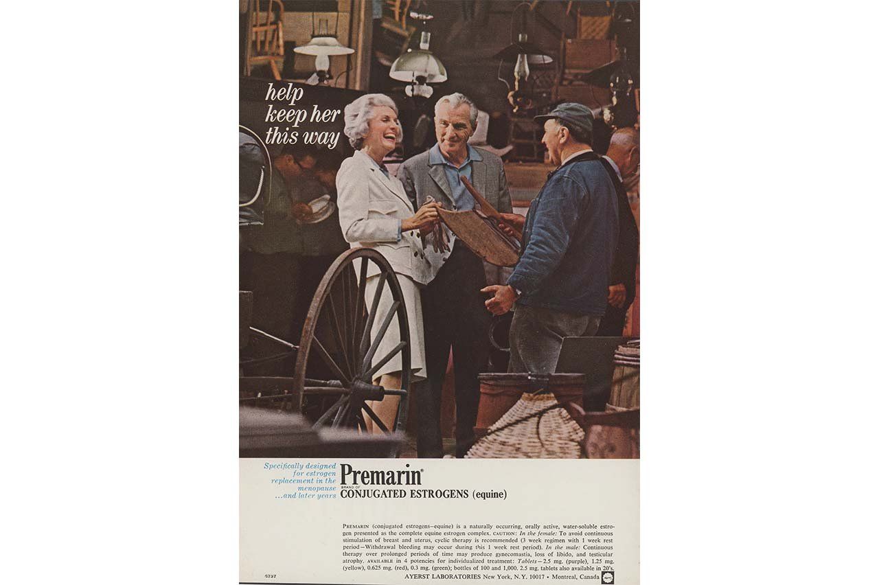 A vintage print advertisement for Premarin (conjugated estrogens). In a warmly lit workshop, an older white woman with gray hair, dressed in a light-colored suit, laughs and smiles while standing beside two older men who are examining a wooden object. Hanging lamps and rustic items fill the background, including a large wagon wheel in the foreground. The ad text reads, “help keep her this way,” suggesting happiness and vitality. Below, smaller text describes Premarin as a hormone therapy for menopause.