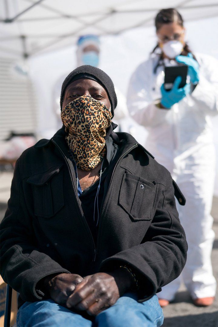 A Black man sits and waits in a leopard print handkerchief over his nose and mouth; a female health care worker in full body PPE works in the background.