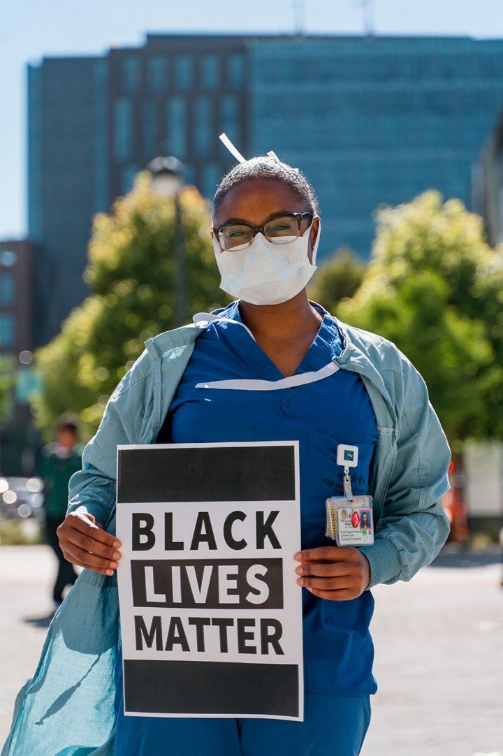 A Black UCSF health care worker stands in front of UCSF Medical Center in scrubs and a face mask, holding a sign that reads “Black Lives Matter.”