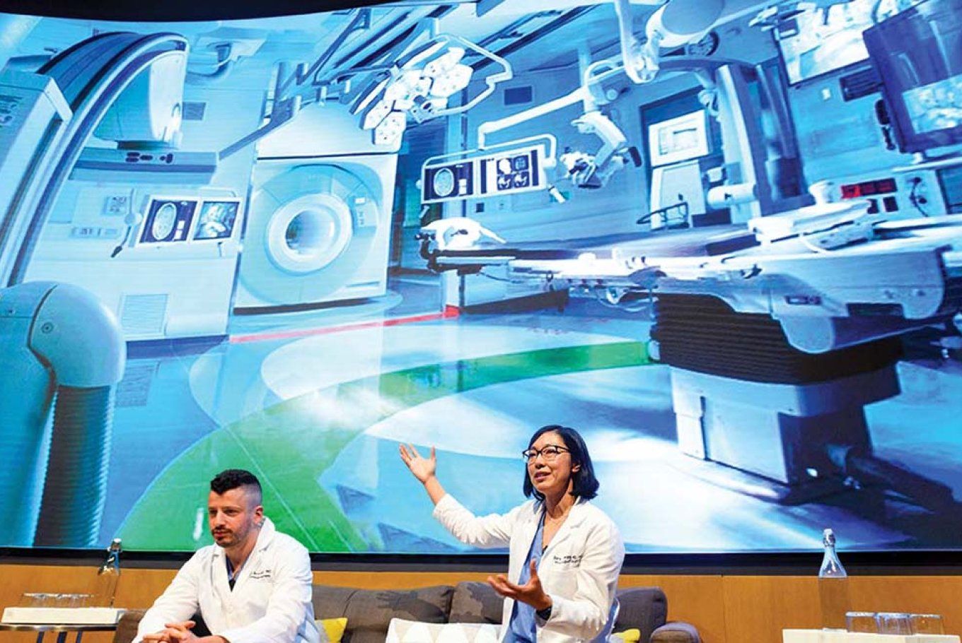 Two surgeons sit in front of a large screen, presenting an operating room of the future.