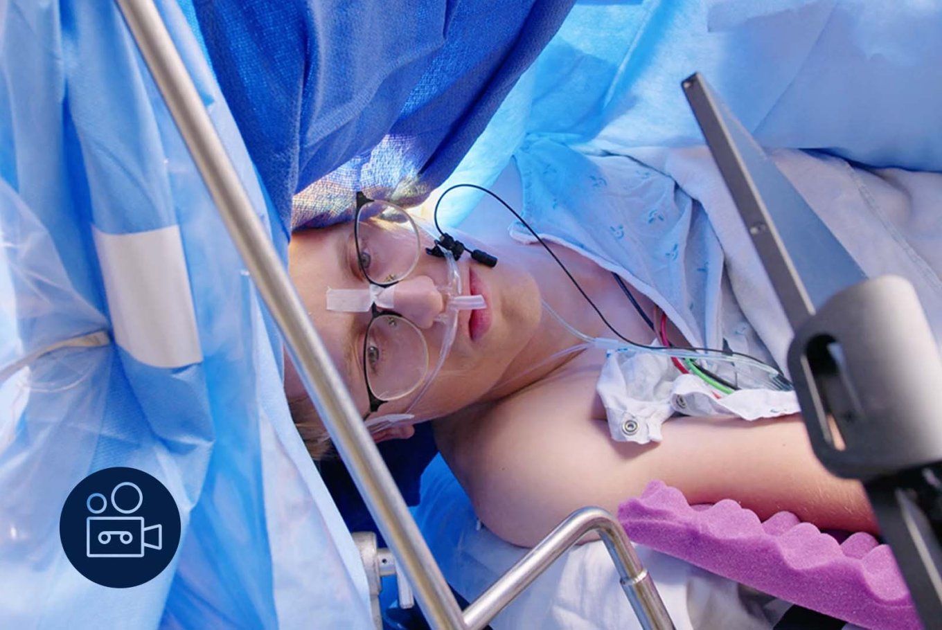 Video icon: A young man in glasses has his eyes open during surgery as he looks at a screen. Blue surgery drapes are all around him.