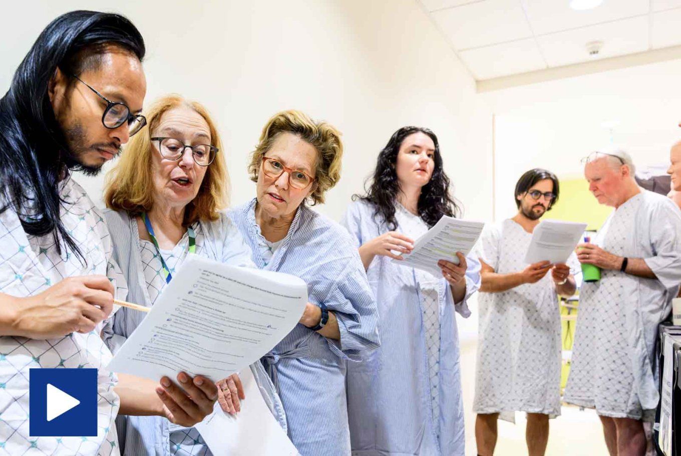 Video play icon: A group of people in hospital gowns stands in a clinical setting, reading and discussing documents while surrounded by medical mannequins.