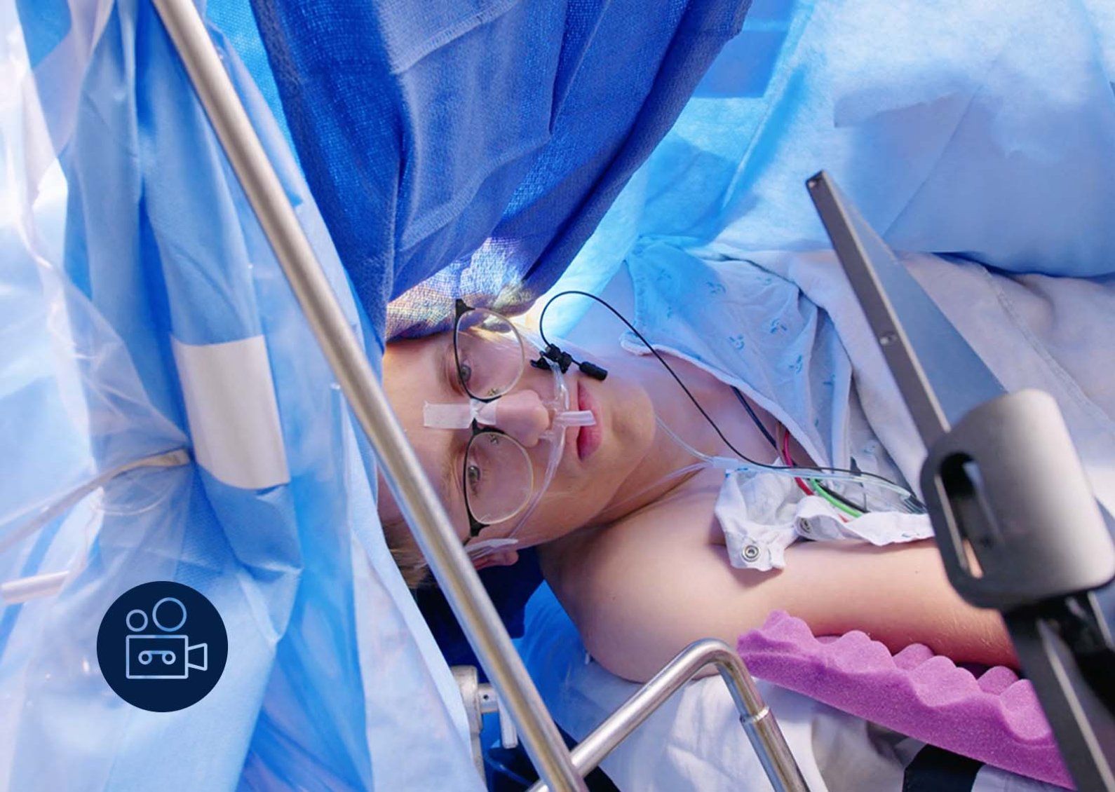 A young man in glasses has his eyes open during surgery as he looks at a screen. Blue surgery drapes are all around him.