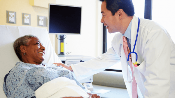 Male doctor with elder female patient by hospital bed. 