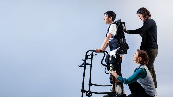 Photo of physical therapist Carolyn Celio, DPT (standing), and rehabilitation aide Cassandra Conlin (kneeling) help Dilan pilot a robotic exoskeleton in front a blank grey background.