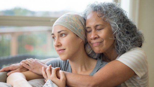 Hawaiian woman in 50s embracing her mid-20s daughter on couch who is fighting cancer.