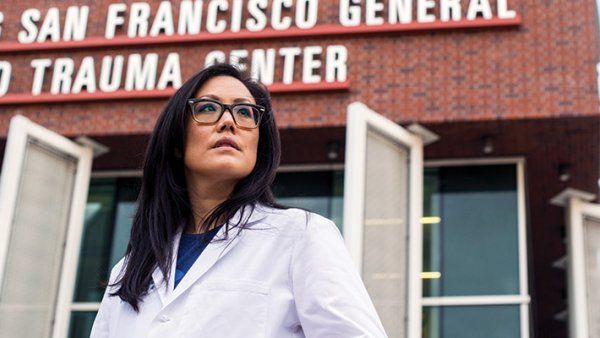 Debbie Yi Madhok wears her doctor’s white coat and stands in front of Zuckerberg San Francisco General Hospital and Trauma Center.
