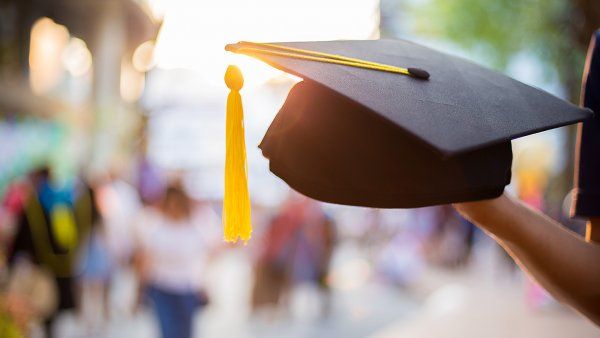 Person holds up a graduation cap with a yellow tassel in front of a busy and blurred street