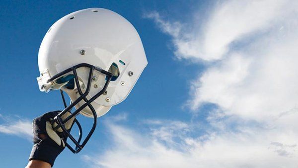 A hand holds up a football helmet in front of a blue sky with clouds.