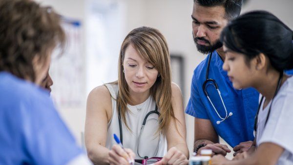 A multi-ethnic group of doctors in an office gathered around a table for a meeting. One woman is taking notes.