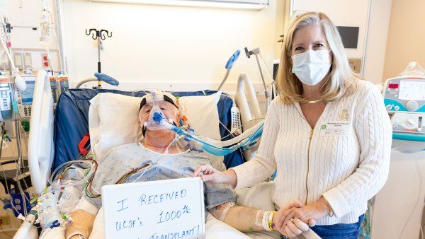Patient Patrick lies in a hospital bed after his lung transplant surgery, with his wife Allison standing beside him. Patrick holds a sign that reads "I received UCSF's 1000th lung transplant."