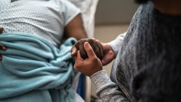 A black patient lies in a hospital bed while their loved one holds their hand