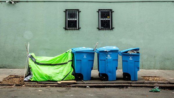 A tent of an unhoused person is set up next to three garbage bins behind a building