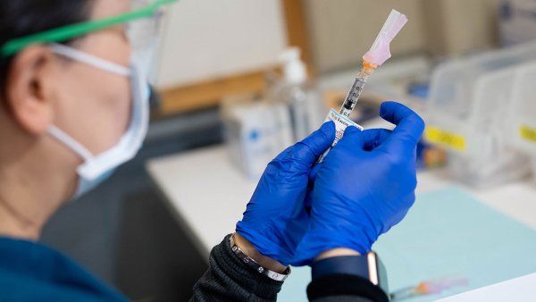 A health professional wearing goggles, a surgical mask, and blue latex gloves prepares a Pfizer COVID-19 vaccine