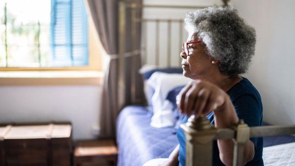 A contemplative elderly Black woman sits on a bed in a brightly lit bedroom and rests her hand on a bedpost