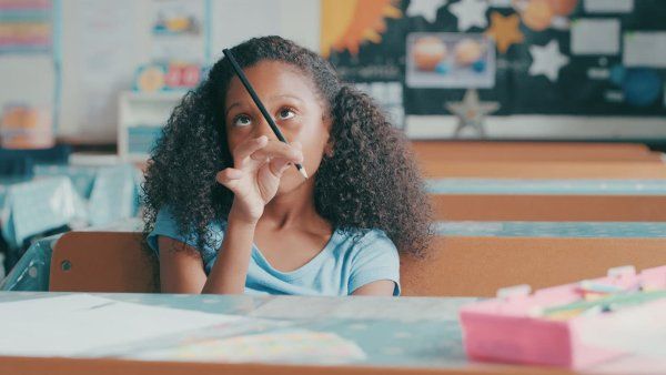 A young Black girl in a classroom stares at her pencil, indicating boredom 