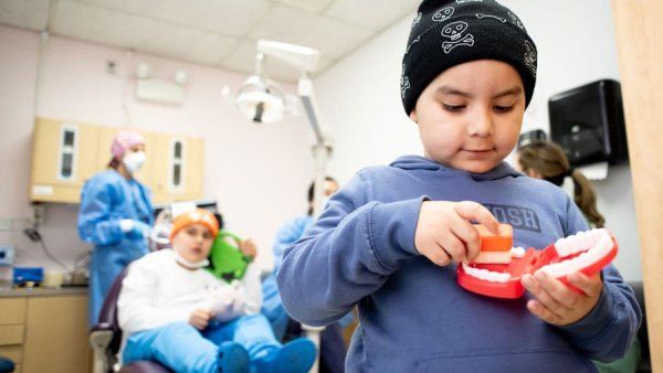 A young boy with a black beanie plays with a dental toy. In the background, a boy wearing an orange beanie sits on a dental chair while a dentist prepares equipment.