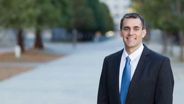 Jon Kleen wears a black suit and blue tie as he poses for a portrait. Trees are in the background.