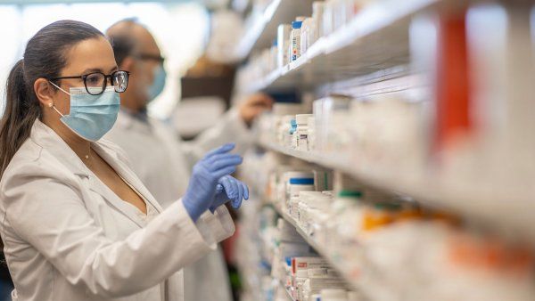 A female pharmacist wearing a lab coat and blue latex gloves picks a bottle of pills from a wall of shelves full of medications.