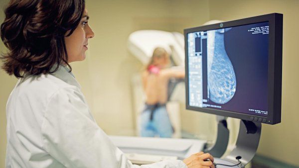 A female doctor examines scans from a mammogram. In the background is a woman standing at the mammogram machine.