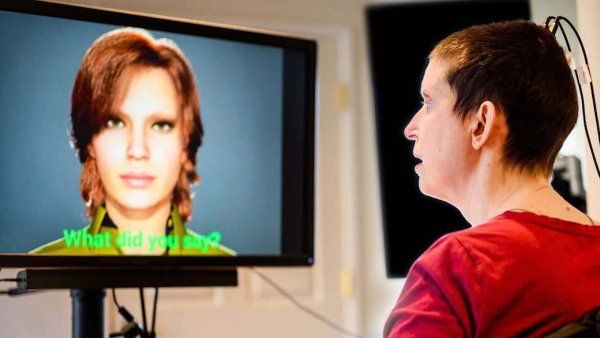 A woman wearing a red shirt and with a buzz haircut sits calmly as an implanted device on her skull is connected to wires that connect to a computer screen. The screen shows an avatar version of the woman and the words "What did you say?"