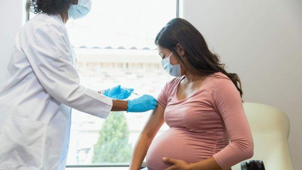 A doctor wearing a white coat and blue latex gloves injects a vaccine into a pregnant woman's arm.