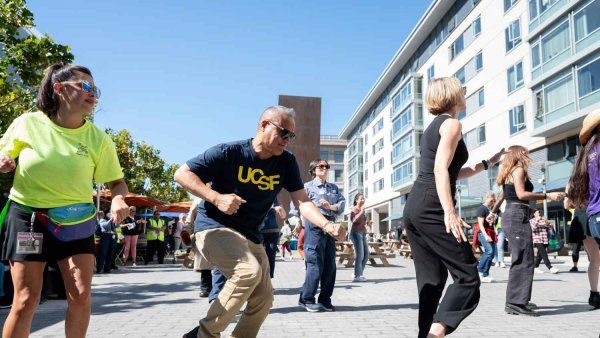 A diverse group of UCSF employees do a line dance outdoors under a bright blue sky.