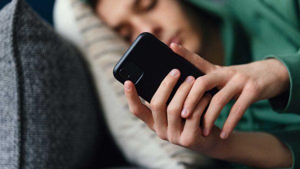 A teenage boy lays on his couch and looks at his phone.