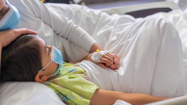 A child and his parent lie together on a hospital bed. They both wear surgial masks.