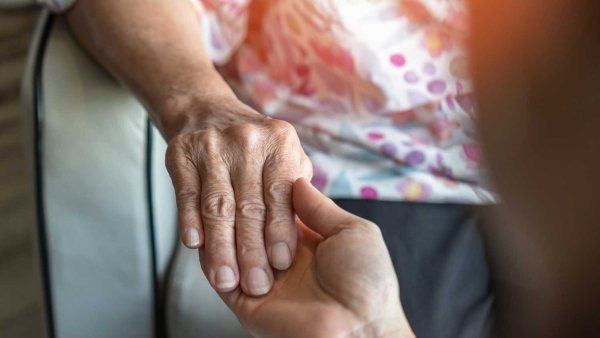 A caregiver holds an elderly woman's hand in comfort.