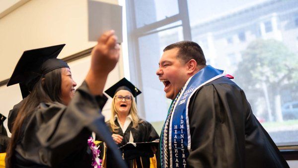 Two new graduates shout joyfully and move in for a hug while outside of a commencement ceremony.